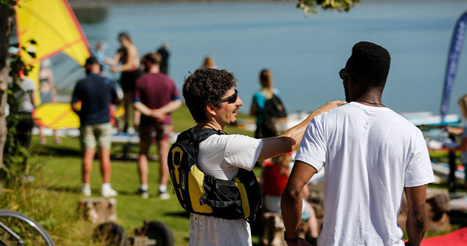 An instructor on shore in a buoyancy aid chatting to someone with other people and windsurfer/dinghy kit and lake in the background.