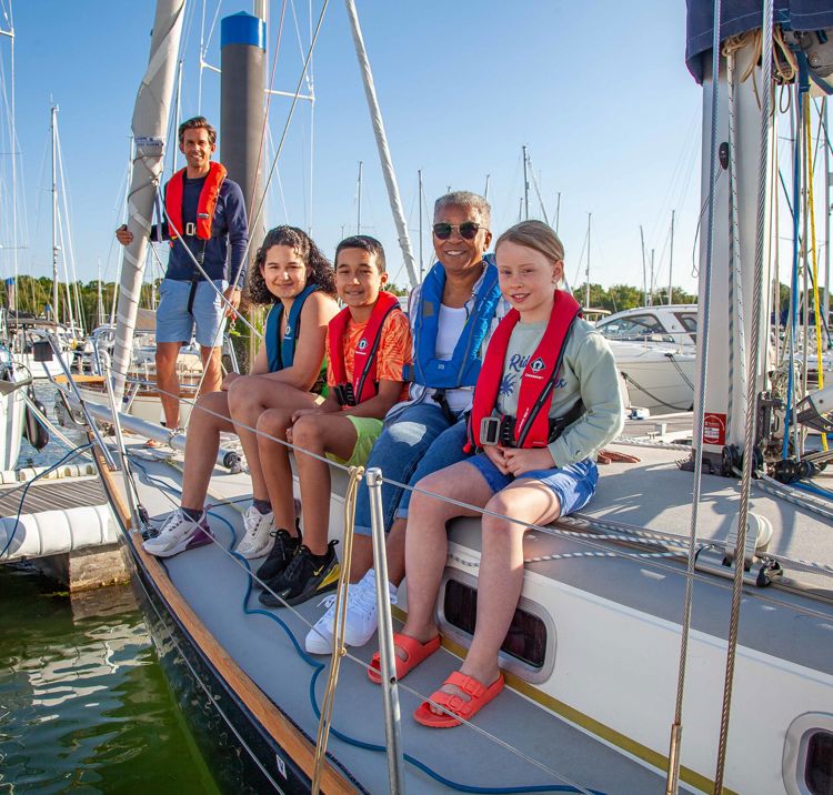 three children and a woman sitting over the side of a yacht whilst it is docked, and man standing behind them